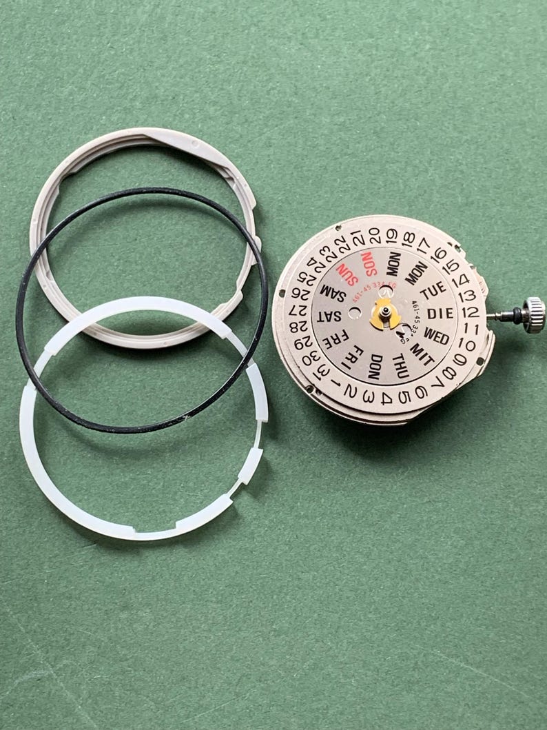May include: Watch movement components, including a dial with day and date displays, and three circular rings. The dial shows days of the week in English and German, and numbers 1-31. The parts are on a green surface.