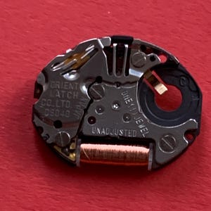 May include: Close-up of a watch movement with the text "ORIENT WATCH CO. LTD D5040" and "ONE (1) JEWEL UNADJUSTED". The movement has silver, gold, and black components on a red background.