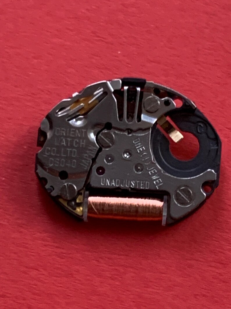 May include: Close-up of a watch movement with the text "ORIENT WATCH CO. LTD D5040" and "ONE (1) JEWEL UNADJUSTED". The movement has silver, gold, and black components on a red background.