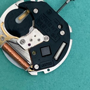 May include: A close-up of an Orient 55910 2J watch movement. The mechanism features a black plastic component with the Orient logo and "Japan" text. The movement includes silver metal components, a copper coil, and a silver screw.