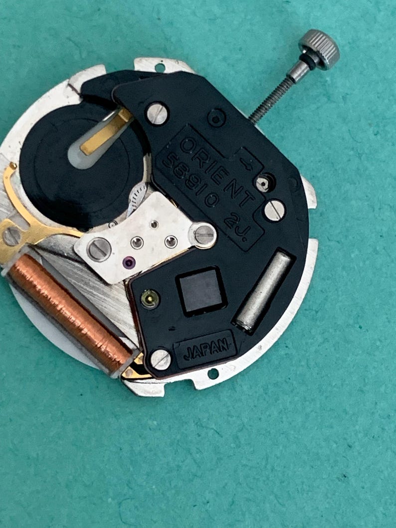 May include: A close-up of an Orient 55910 2J watch movement. The mechanism features a black plastic component with the Orient logo and "Japan" text. The movement includes silver metal components, a copper coil, and a silver screw.