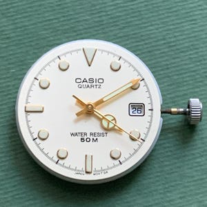 K&ouml;nnte beinhalten: Ein Casio Quartz Zifferblatt mit cremefarbenem Zifferblatt, goldfarbenen Zeigern und Markierungen sowie einem Datumsfenster mit der Zahl 26. Das Zifferblatt tr&auml;gt die Aufschrift "CASIO QUARTZ" und "WATER RESIST 50M".