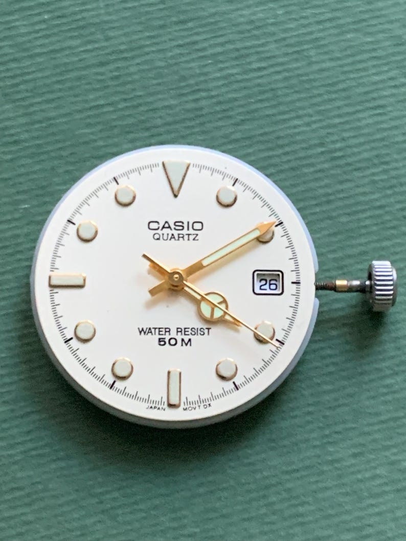 K&ouml;nnte beinhalten: Ein Casio Quartz Zifferblatt mit cremefarbenem Zifferblatt, goldfarbenen Zeigern und Markierungen sowie einem Datumsfenster mit der Zahl 26. Das Zifferblatt tr&auml;gt die Aufschrift "CASIO QUARTZ" und "WATER RESIST 50M".