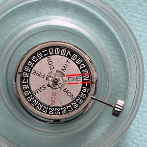 May include: Close-up of a watch movement with a silver and black dial. The dial displays the days of the week and numbers 1-31. The word "DIE" is visible. A clear plastic case surrounds the watch movement.