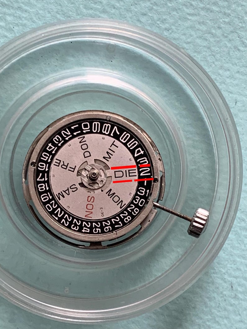 May include: Close-up of a watch movement with a silver and black dial. The dial displays the days of the week and numbers 1-31. The word "DIE" is visible. A clear plastic case surrounds the watch movement.