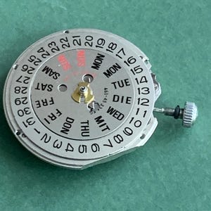 May include: A silver watch movement with a day and date display. The dial has black numbers for the date and days of the week in multiple languages. A small gold component is in the center. The watch mechanism is on a green surface.