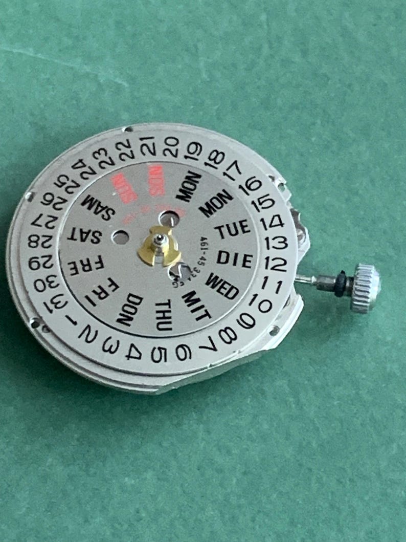 May include: A silver watch movement with a day and date display. The dial has black numbers for the date and days of the week in multiple languages. A small gold component is in the center. The watch mechanism is on a green surface.