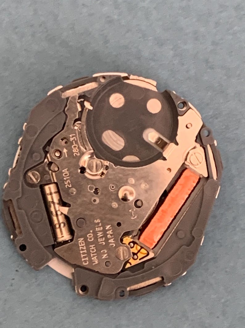 May include: A close-up of a Citizen watch movement, featuring intricate gears, circuits, and a battery. The movement is primarily silver and gray, with a bright orange component and the text "CITIZEN WATCH CO. NJ JEWELS JAPAN".