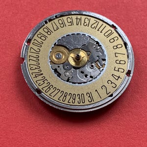 May include: A watch movement with a gold-colored dial and silver-colored metal casing. The dial features the numbers 1 through 31, indicating the days of the month. The intricate gears and mechanisms are visible within the watch movement.