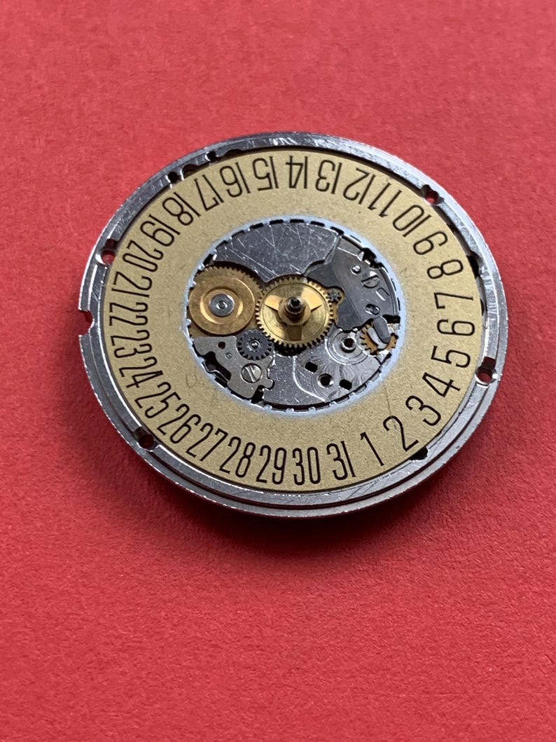 May include: A watch movement with a gold-colored dial and silver-colored metal casing. The dial features the numbers 1 through 31, indicating the days of the month. The intricate gears and mechanisms are visible within the watch movement.