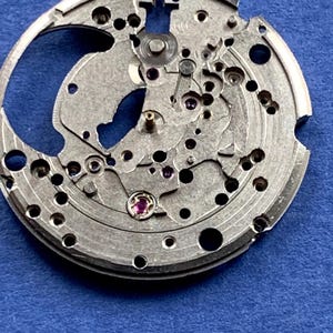 Puede incluir: Primer plano de un movimiento de reloj plateado con intrincados engranajes, agujeros y peque&ntilde;as joyas. Los componentes mec&aacute;nicos est&aacute;n dispuestos en un patr&oacute;n circular, mostrando la precisi&oacute;n del funcionamiento interno del reloj. El fondo es azul liso.