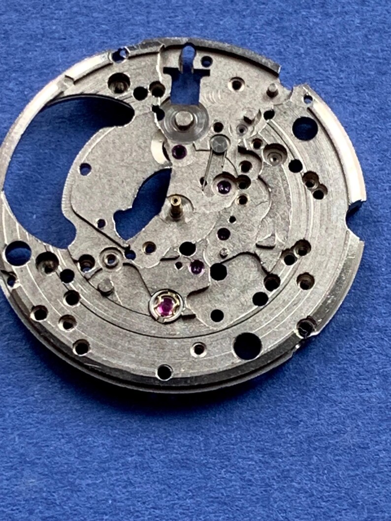 Puede incluir: Primer plano de un movimiento de reloj plateado con intrincados engranajes, agujeros y peque&ntilde;as joyas. Los componentes mec&aacute;nicos est&aacute;n dispuestos en un patr&oacute;n circular, mostrando la precisi&oacute;n del funcionamiento interno del reloj. El fondo es azul liso.