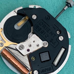 May include: A detailed view of a watch movement, displaying the text "ORIENT 56910 2J" and "JAPAN". The mechanism includes a black plate, silver metal parts, and a copper coil. A small silver screw is also visible.