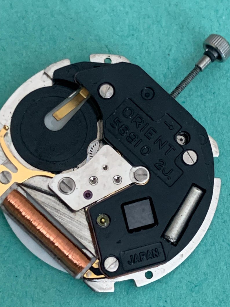 May include: A detailed view of a watch movement, displaying the text "ORIENT 56910 2J" and "JAPAN". The mechanism includes a black plate, silver metal parts, and a copper coil. A small silver screw is also visible.