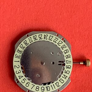 K&ouml;nnte beinhalten: Ein Uhrwerk mit silberfarbener Metallbasis und einem kreisf&ouml;rmigen Datumszifferblatt. Das Zifferblatt zeigt die Zahlen 1 bis 31 in hellgr&uuml;ner Schrift. Auf der rechten Seite ist eine kleine goldfarbene Krone zu sehen.