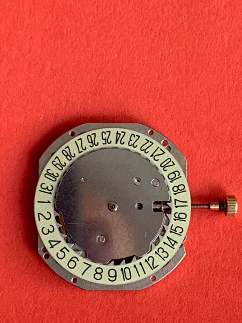 K&ouml;nnte beinhalten: Ein Uhrwerk mit silberfarbener Metallbasis und einem kreisf&ouml;rmigen Datumszifferblatt. Das Zifferblatt zeigt die Zahlen 1 bis 31 in hellgr&uuml;ner Schrift. Auf der rechten Seite ist eine kleine goldfarbene Krone zu sehen.