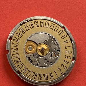 May include: A close-up of a watch movement with a gold-colored date wheel. The wheel has black numbers from 1 to 31. The movement's gears and components are visible, set against a red background.