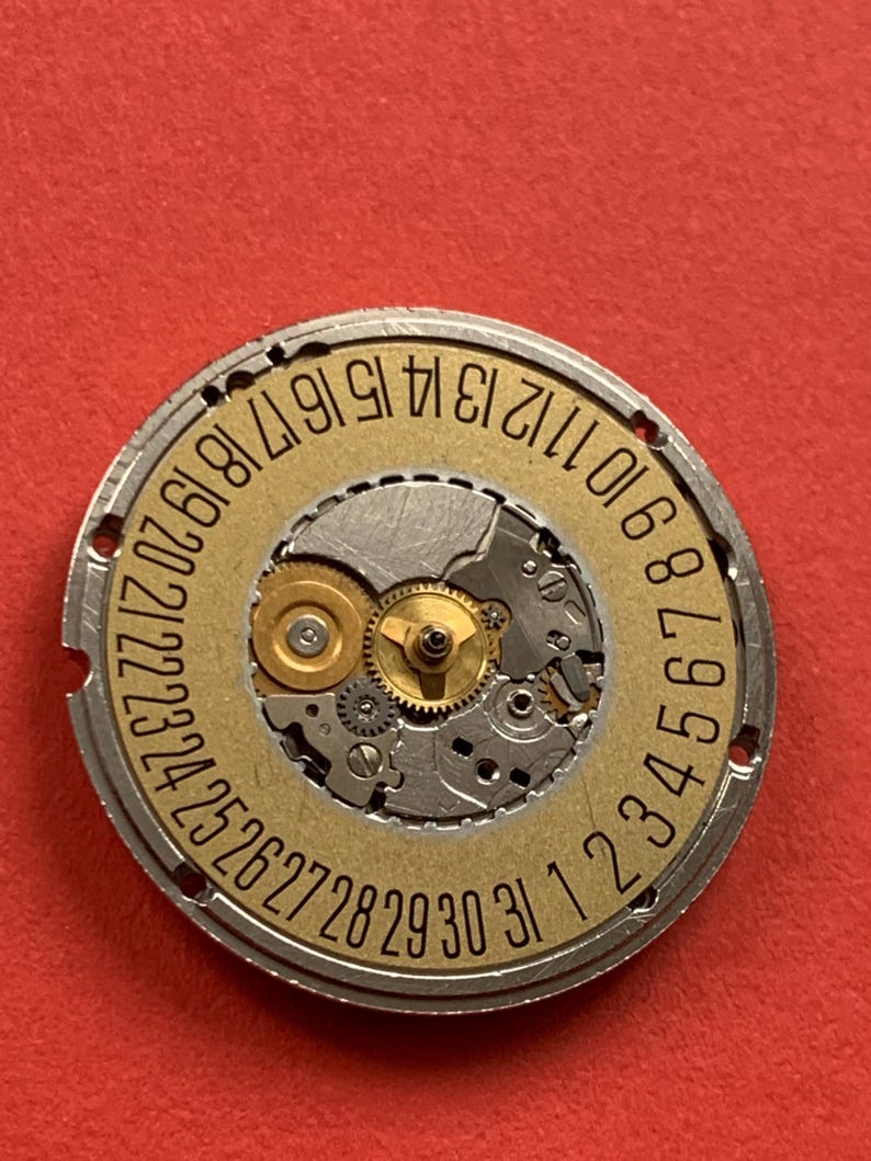 May include: A close-up of a watch movement with a gold-colored date wheel. The wheel has black numbers from 1 to 31. The movement's gears and components are visible, set against a red background.