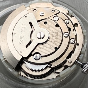 May include: Close-up of a Seiko watch movement. The silver-toned mechanism features multiple gears, screws, and the brand name "SEIKO" printed on a circular component. The movement is set against a clear background.