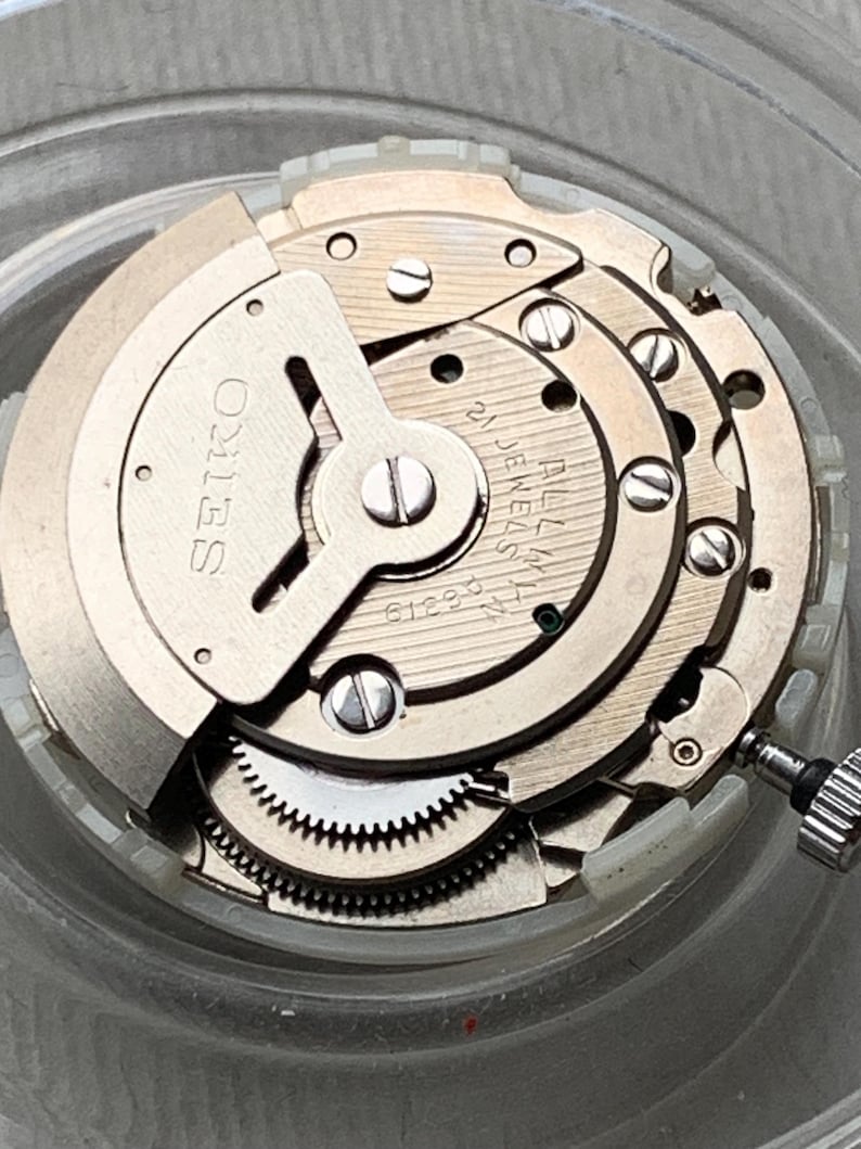 May include: Close-up of a Seiko watch movement. The silver-toned mechanism features multiple gears, screws, and the brand name "SEIKO" printed on a circular component. The movement is set against a clear background.