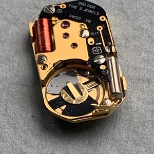 May include: Close-up of a watch movement with gold-colored components, including a coil, gears, and a battery. The black circuit board has the text "280 002 FIVE 5 JEWELS SWISS V8". The watch movement is on a gray surface.