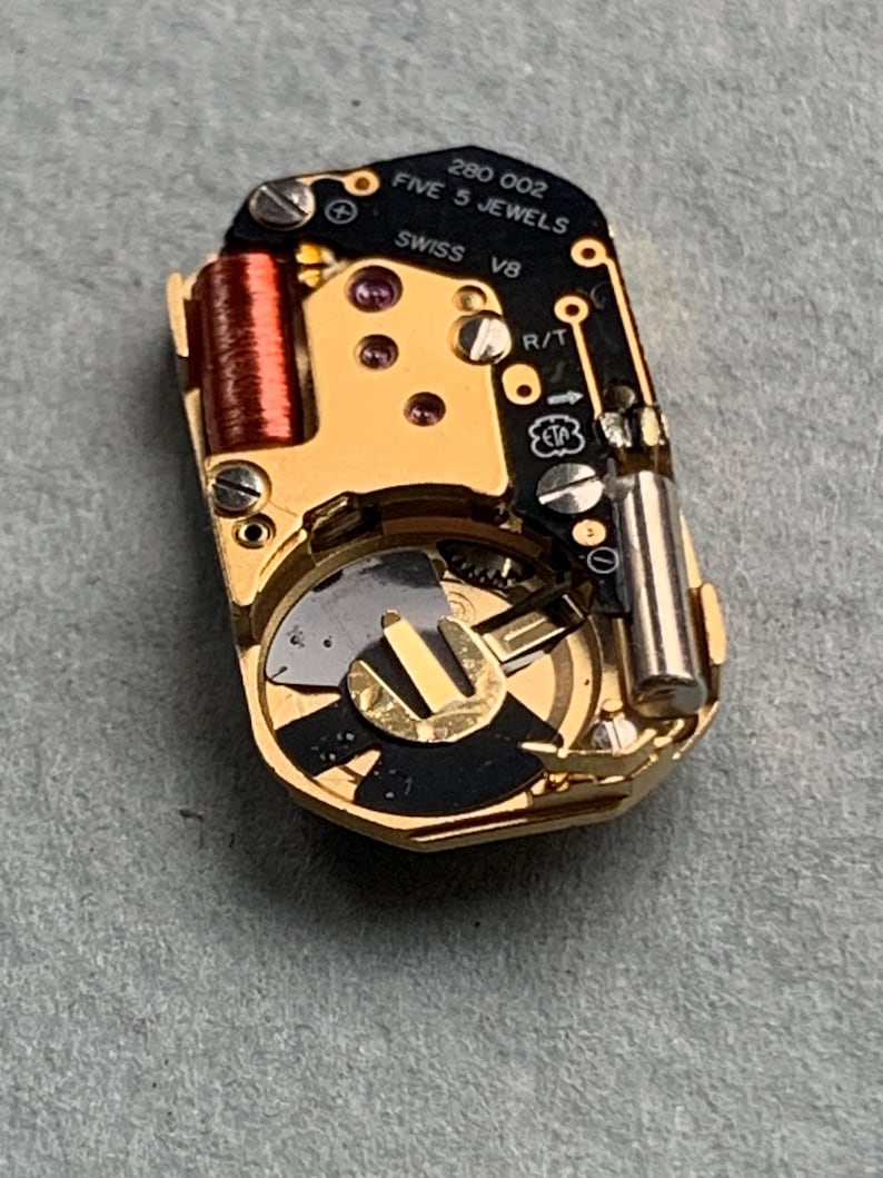 May include: Close-up of a watch movement with gold-colored components, including a coil, gears, and a battery. The black circuit board has the text "280 002 FIVE 5 JEWELS SWISS V8". The watch movement is on a gray surface.