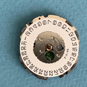 May include: A watch movement with a white date wheel featuring black numbers from 1 to 31. The movement has silver-colored gears and a green component. The text "E6-3-3" is visible.