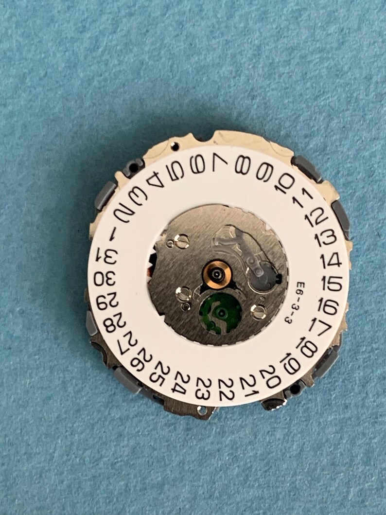 May include: A watch movement with a white date wheel featuring black numbers from 1 to 31. The movement has silver-colored gears and a green component. The text "E6-3-3" is visible.
