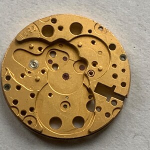 May include: Close-up of a gold-colored watch movement with intricate gears, holes, and small red jewels. The number "2571" is stamped on the edge. The watch mechanism is circular and detailed.