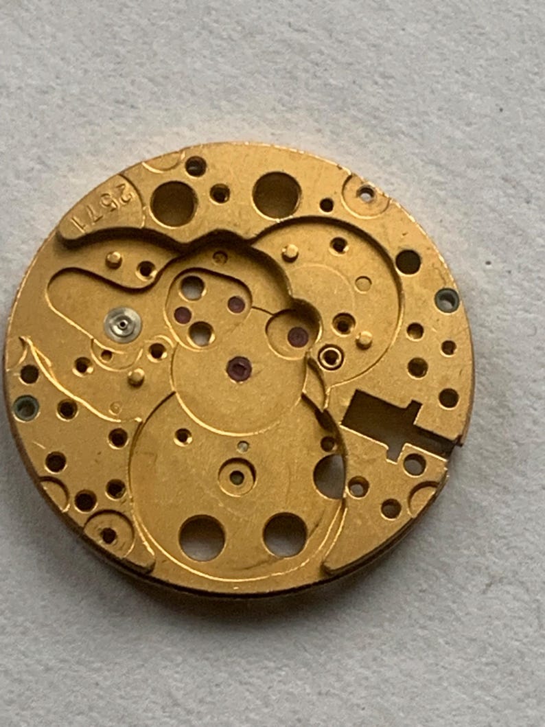 May include: Close-up of a gold-colored watch movement with intricate gears, holes, and small red jewels. The number "2571" is stamped on the edge. The watch mechanism is circular and detailed.