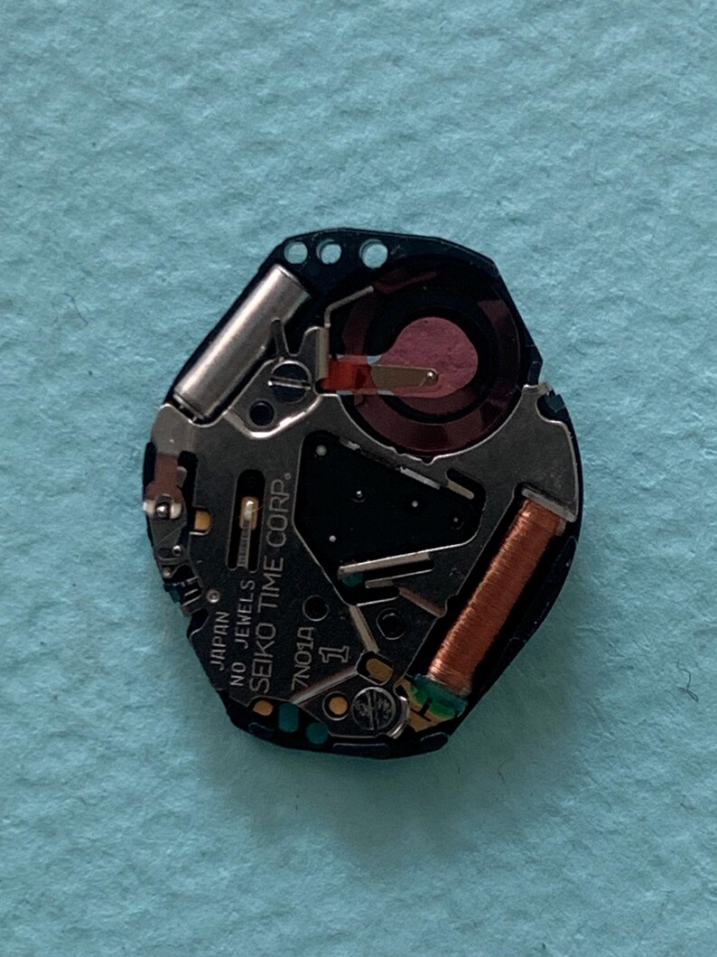May include: A detailed view of a Seiko watch movement, showcasing its internal mechanisms. The movement is predominantly black, with silver and copper-toned elements. The text "SEIKO TIME CORP" and "JAPAN NO JEWELS" are printed on the movement.