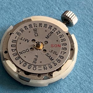 May include: Close-up of a watch movement with a silver-colored dial and a white plastic ring. The dial displays the days of the week in German and numbers for the date. A silver crown is attached to the side.