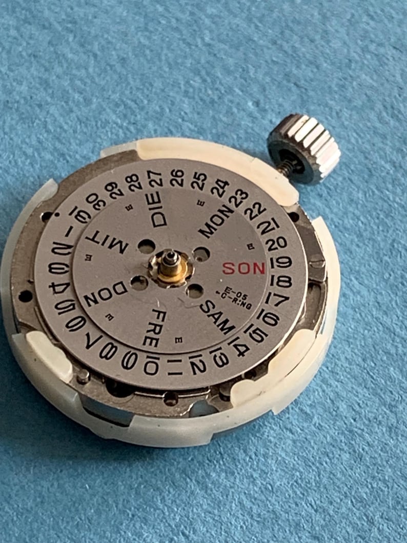 May include: Close-up of a watch movement with a silver-colored dial and a white plastic ring. The dial displays the days of the week in German and numbers for the date. A silver crown is attached to the side.