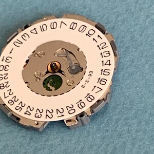 May include: A close-up of a watch movement component. The component features a white circular dial with black numbers from 1 to 31, a silver metal frame, and internal gears. The background is a light blue surface.