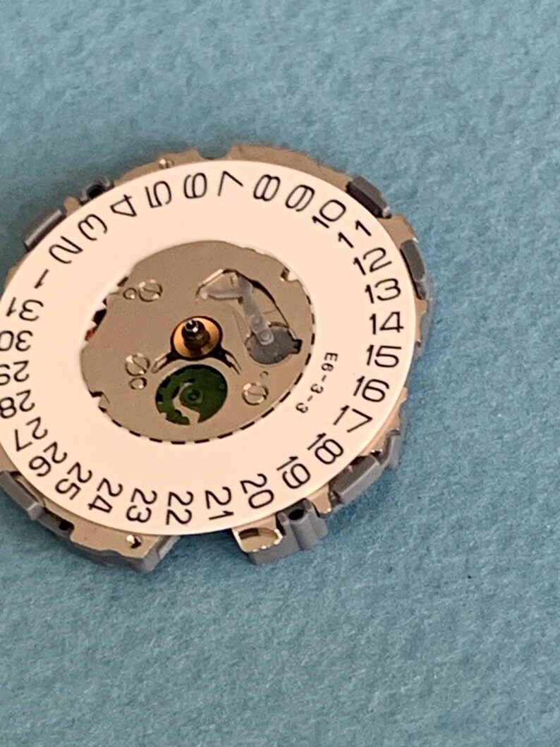 May include: A close-up of a watch movement component. The component features a white circular dial with black numbers from 1 to 31, a silver metal frame, and internal gears. The background is a light blue surface.
