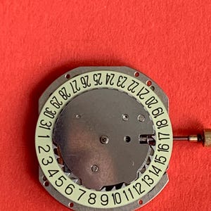K&ouml;nnte beinhalten: Ein Uhrwerk mit silberfarbenem Zifferblatt und einem wei&szlig;en Ring mit den Monatstagen von 1 bis 31. Eine kleine goldfarbene Schraube befindet sich auf der rechten Seite. Der Hintergrund ist einfarbig rot.