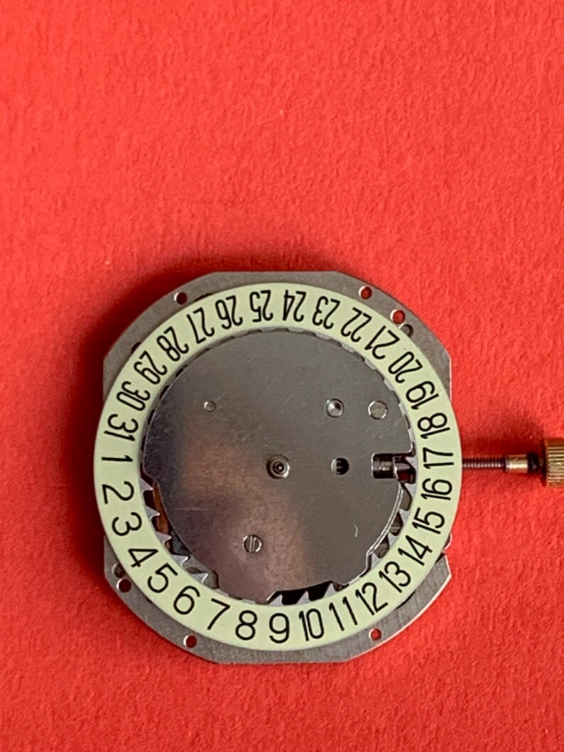 K&ouml;nnte beinhalten: Ein Uhrwerk mit silberfarbenem Zifferblatt und einem wei&szlig;en Ring mit den Monatstagen von 1 bis 31. Eine kleine goldfarbene Schraube befindet sich auf der rechten Seite. Der Hintergrund ist einfarbig rot.
