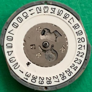 May include: A watch movement with a white dial featuring black numerals from 1 to 31, indicating the days of the month. The dial is surrounded by a gray ring. The internal mechanism is visible, with silver-colored gears and components.