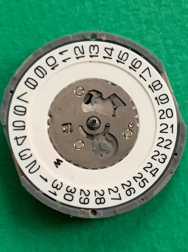 May include: A watch movement with a white dial featuring black numerals from 1 to 31, indicating the days of the month. The dial is surrounded by a gray ring. The internal mechanism is visible, with silver-colored gears and components.