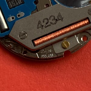 May include: Close-up of a watch movement with a blue circuit board and metallic components. The number "4234" is etched on a silver plate, with a copper coil visible. Additional markings include "B151 0402" and "955 411".