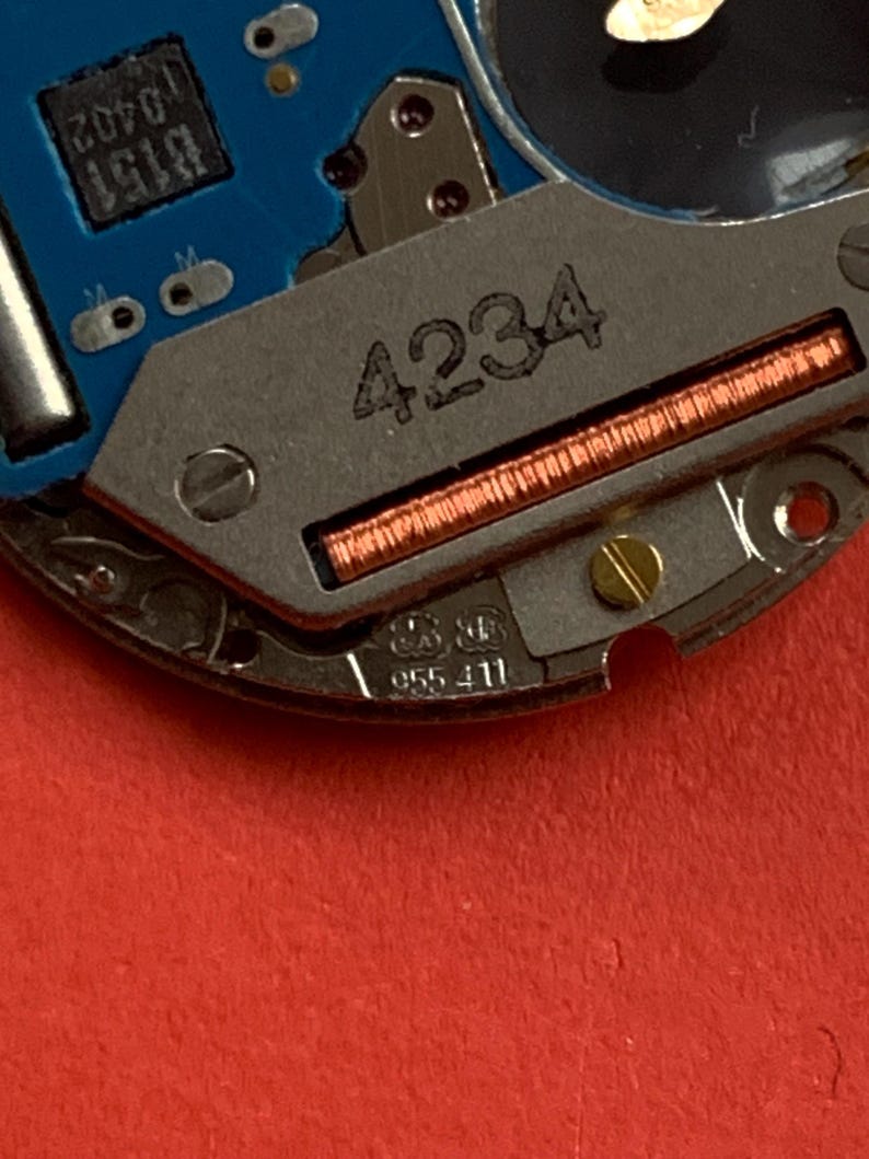 May include: Close-up of a watch movement with a blue circuit board and metallic components. The number "4234" is etched on a silver plate, with a copper coil visible. Additional markings include "B151 0402" and "955 411".