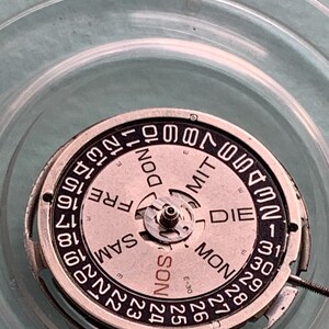 May include: Close-up of a watch movement with a circular dial. The dial features the days of the week in German and numbers for the date. The watch mechanism is silver-toned with a light pink dial and black text.