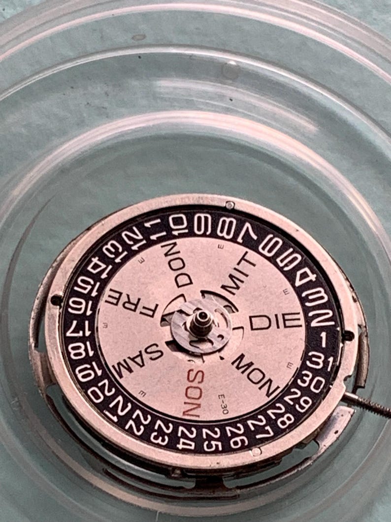 May include: Close-up of a watch movement with a circular dial. The dial features the days of the week in German and numbers for the date. The watch mechanism is silver-toned with a light pink dial and black text.