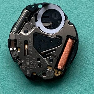 May include: A close-up of a watch movement with various components. The watch movement is black and silver, with a copper coil and the text "SEIKO TIME CORP." and "JAPAN" visible. The background is a light teal color.
