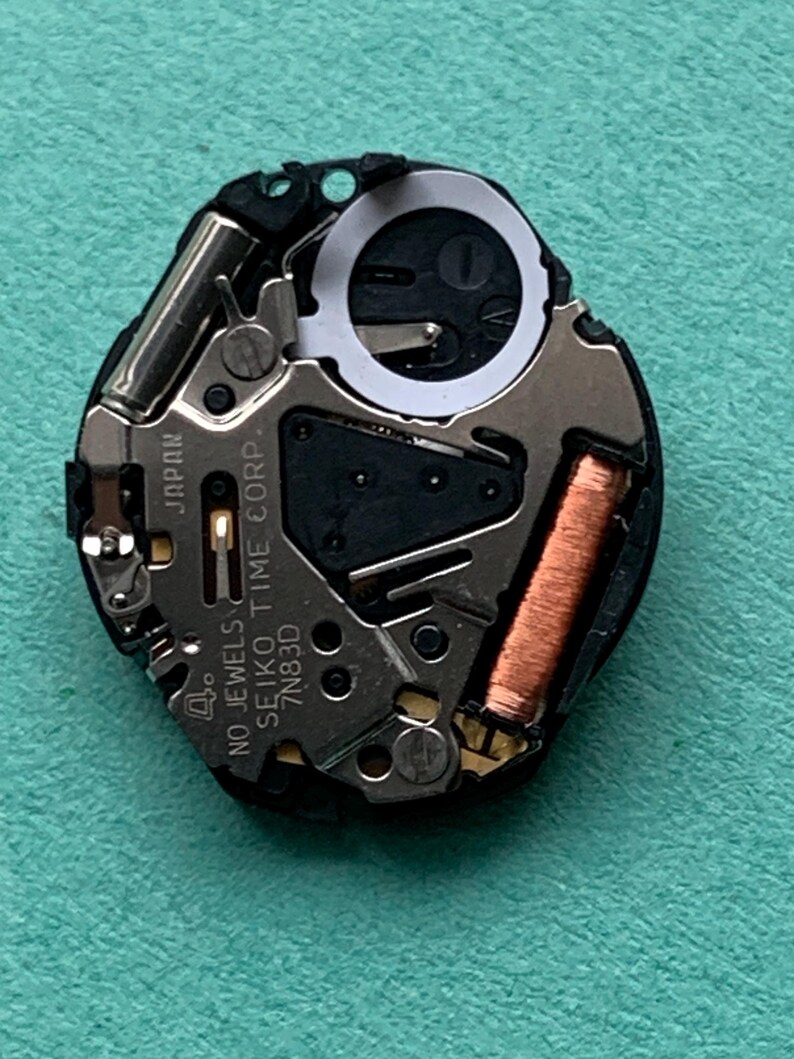 May include: A close-up of a watch movement with various components. The watch movement is black and silver, with a copper coil and the text "SEIKO TIME CORP." and "JAPAN" visible. The background is a light teal color.