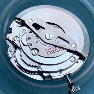 May include: Close-up of a Seiko watch movement. The watch mechanism features silver-coloured metal components, including gears, screws, and a balance wheel. The text "SEIKO JEWELS" is visible on the watch face. The watch movement is set inside a clear container.