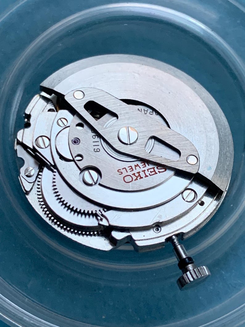 May include: Close-up of a Seiko watch movement. The watch mechanism features silver-coloured metal components, including gears, screws, and a balance wheel. The text "SEIKO JEWELS" is visible on the watch face. The watch movement is set inside a clear container.