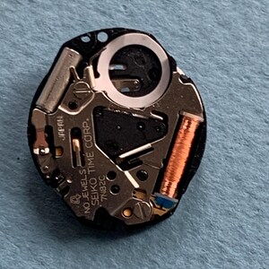 K&ouml;nnte beinhalten: Nahaufnahme eines Uhrwerks mit dem Text "NO JEWELS SEIKO TIME CORP. JAPAN 7N82C". Das Uhrwerk verf&uuml;gt &uuml;ber silberfarbene Metallkomponenten, eine kupferfarbene Spule und eine schwarze Basis. Das Uhrwerk liegt auf einer blauen Oberfl&auml;che.
