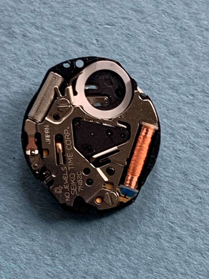 K&ouml;nnte beinhalten: Nahaufnahme eines Uhrwerks mit dem Text "NO JEWELS SEIKO TIME CORP. JAPAN 7N82C". Das Uhrwerk verf&uuml;gt &uuml;ber silberfarbene Metallkomponenten, eine kupferfarbene Spule und eine schwarze Basis. Das Uhrwerk liegt auf einer blauen Oberfl&auml;che.