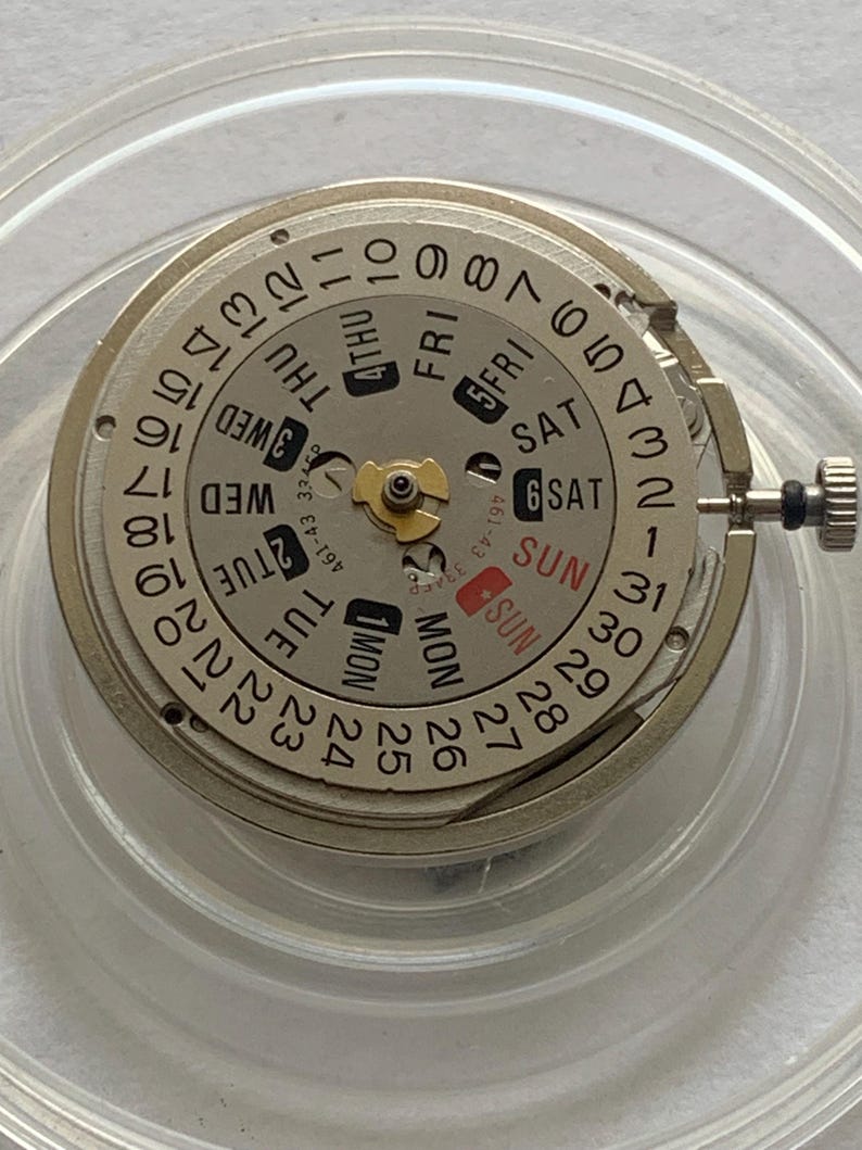 May include: A watch movement with a silver-colored dial and a day-date display. The dial features numbers, day abbreviations (Mon, Tue, Wed, Thu, Fri, Sat, Sun), and a small crown. The movement is set in a clear plastic holder.